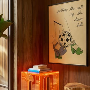 May include: An orange acrylic side table with a stack of books sits next to a framed illustration. The illustration features a disco ball and the text "follow the call of the disco ball". The room has wood paneling.