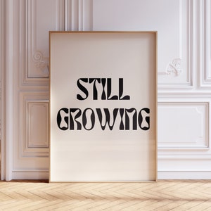 K&ouml;nnte beinhalten: Ein gerahmter Druck mit wei&szlig;em Hintergrund und schwarzem Text, der "STILL GROWING" in einer fetten, stilisierten Schriftart lautet.