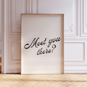 May include: A framed print with the text "Meet you there?" in a stylish script font. The print has a light background and a gold frame. The print is leaning against a white wall with decorative molding, and the floor is made of wood.