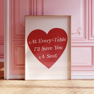 以下が含まれることがあります： 赤いハートの形をしたフレーム付きプリントで、白い文字で「At Every Table I'll Save You A Seat」と書かれています。プリントは白い背景で、金色のフレームで囲まれています。