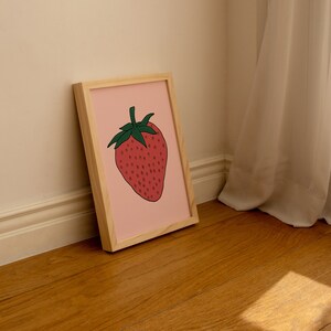 May include: A framed illustration of a red strawberry with green leaves on a pink background. The frame is made of light wood.