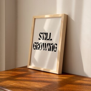 K&ouml;nnte beinhalten: Ein gerahmter Druck mit dem Text "STILL GROWING" in einer schwarzen, stilisierten Schrift auf wei&szlig;em Hintergrund. Der Rahmen ist aus hellem Holz.