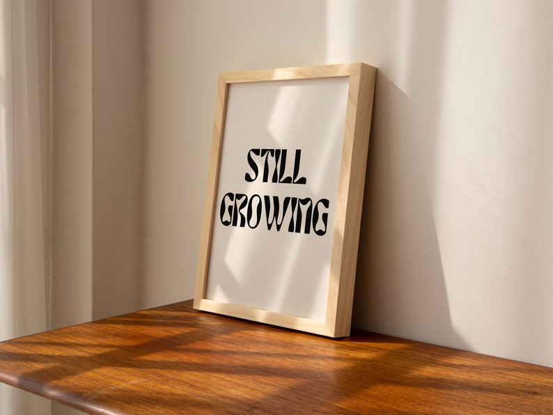 K&ouml;nnte beinhalten: Ein gerahmter Druck mit dem Text "STILL GROWING" in einer schwarzen, stilisierten Schrift auf wei&szlig;em Hintergrund. Der Rahmen ist aus hellem Holz.