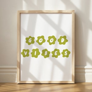 May include: A framed print with the words "GOOD VIBES" in white letters on a green, flower-shaped background. The print is in a light wood frame and is displayed against a white wall.