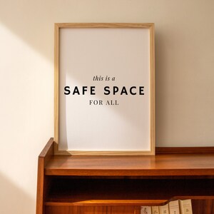 May include: A framed print with the text "this is a SAFE SPACE FOR ALL" in black lettering on a white background. The frame is made of light wood and is sitting on a wooden shelf.