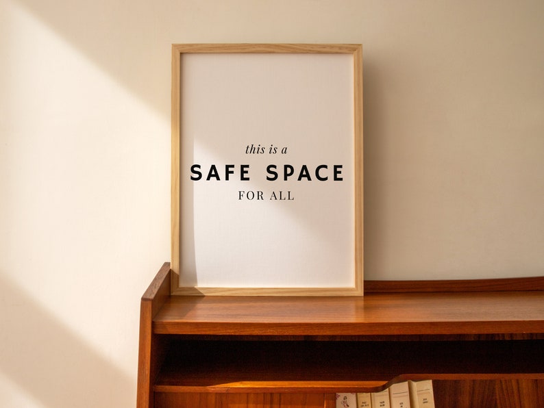 May include: A framed print with the text "this is a SAFE SPACE FOR ALL" in black lettering on a white background. The frame is made of light wood and is sitting on a wooden shelf.