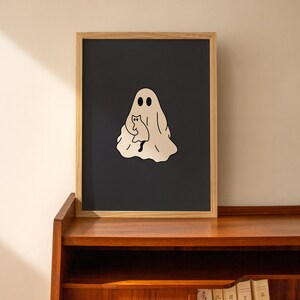 May include: A framed print with a black background and a white outline of a ghost holding a cat. The ghost has two eyes and a simple smile.