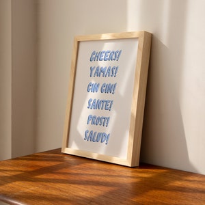 May include: A framed print with the words "CHEERS! YAMAS! CIN CIN! SANTE! PROST! SALUD!" in blue text on a white background.