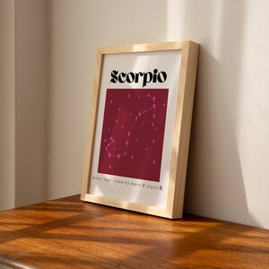 May include: A framed print with a burgundy background and a pink constellation of the Scorpio zodiac sign. The text "Scorpio" is in black bold letters at the top of the print. The text "water sign, ruled by mars & pluto" is at the bottom of the print.