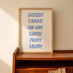May include: A framed print with the words "CHEERS! YAMAS! CIN CIN! SANTE! PROST! SALUD!" in blue text on a white background.