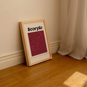 May include: A framed print with a burgundy background and a pink constellation of the Scorpio zodiac sign. The text "Scorpio" is in black bold letters at the top of the print. The text "water sign, ruled by mars & pluto" is at the bottom of the print.