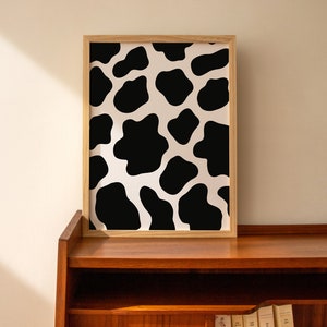 May include: A framed print with a black and white cow print pattern. The print is in a light wood frame and is hanging on a wall.