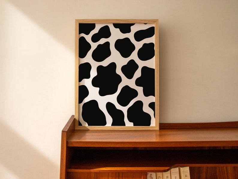 May include: A framed print with a black and white cow print pattern. The print is in a light wood frame and is hanging on a wall.