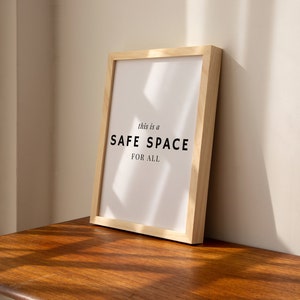 May include: A framed print with the text "This is a SAFE SPACE FOR ALL" in black lettering on a white background. The frame is made of light wood and is sitting on a wooden surface.