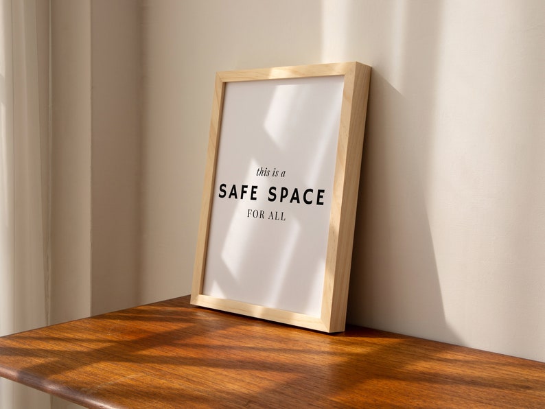 May include: A framed print with the text "This is a SAFE SPACE FOR ALL" in black lettering on a white background. The frame is made of light wood and is sitting on a wooden surface.
