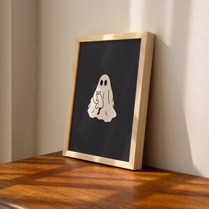 May include: A framed black and white illustration of a ghost holding a cat. The ghost has two eyes and a simple smile. The cat is sitting upright with its tail curled around its body. The illustration is on a black background and is framed in a light wood frame.