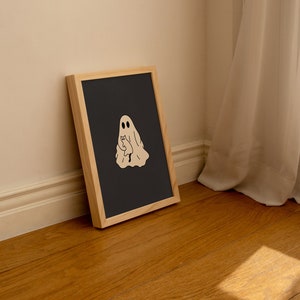 May include: A framed black and white illustration of a ghost holding a cat. The ghost is white and has two eyes. The cat is brown and has a white chest. The frame is made of wood and has a light brown finish.