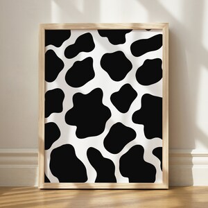 May include: A framed print with a black and white cow print pattern. The print is in a light wood frame.