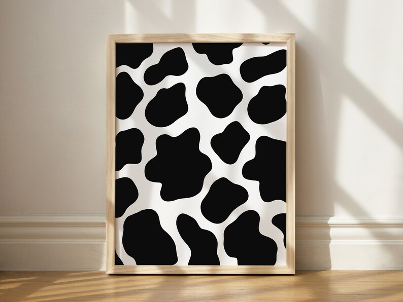 May include: A framed print with a black and white cow print pattern. The print is in a light wood frame.