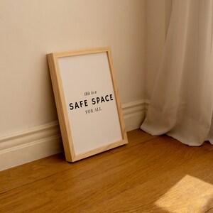 May include: A wooden framed print with the text "This is a SAFE SPACE FOR ALL" on a white background. The print is leaning against a wall with a white curtain in the background.