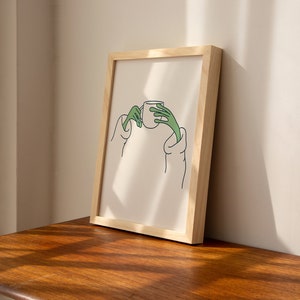 May include: A framed illustration of a person holding a cup with green hands. The illustration is on a white background and is framed in a natural wood frame.