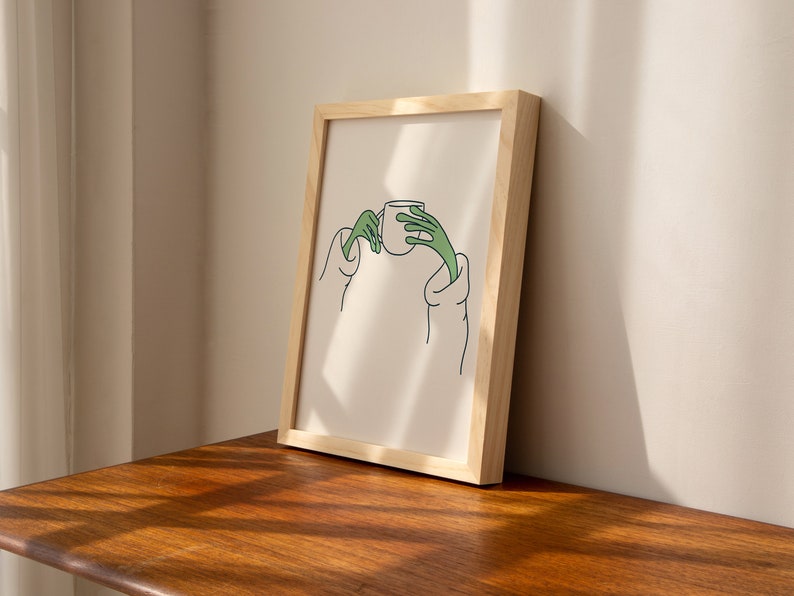 May include: A framed illustration of a person holding a cup with green hands. The illustration is on a white background and is framed in a natural wood frame.