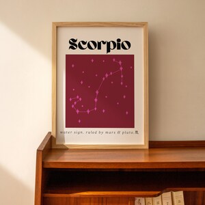 May include: A framed print with a burgundy background and a pink constellation of the zodiac sign Scorpio. The text "Scorpio" is in black bold font and the text "water sign. ruled by mars & pluto." is in smaller black font.