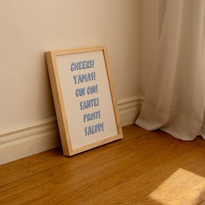 May include: A framed print with the words "CHEERS! YAMAS! CIN CIN! SANTE! PROST! SALUD!" in blue lettering on a white background.