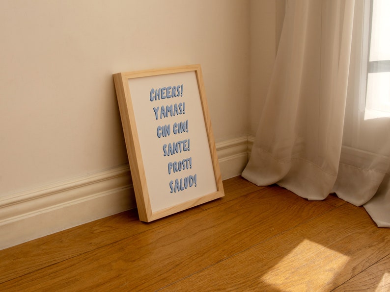 May include: A framed print with the words "CHEERS! YAMAS! CIN CIN! SANTE! PROST! SALUD!" in blue lettering on a white background.