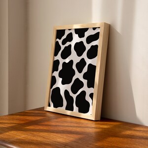 May include: A framed print with a black and white cow print pattern. The frame is made of natural wood.