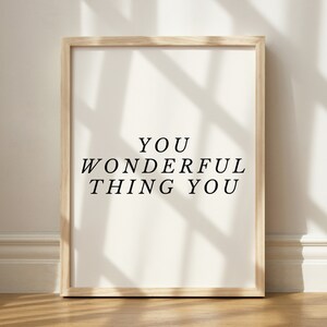Op de afbeelding: Een ingelijst print met de tekst "YOU WONDERFUL THING YOU" in zwarte letters op een witte achtergrond. De lijst is van licht hout en de print hangt aan een witte muur.