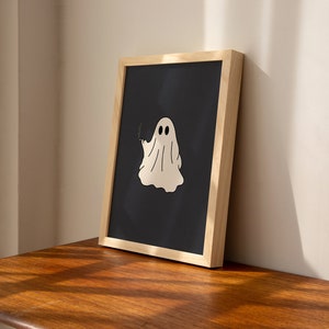 May include: A framed print of a white ghost with two eyes on a black background. The frame is made of light wood.
