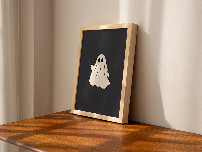 May include: A framed print of a white ghost with two eyes on a black background. The frame is made of light wood.