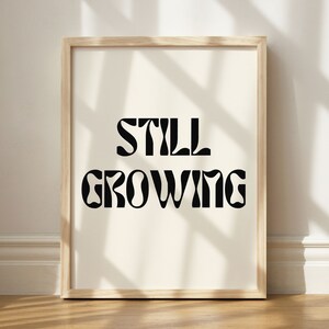 K&ouml;nnte beinhalten: Ein gerahmter Druck mit dem Text "STILL GROWING" in einer Retro-Schriftart. Der Druck ist auf wei&szlig;em Hintergrund und in einem hellen Holzrahmen gerahmt.