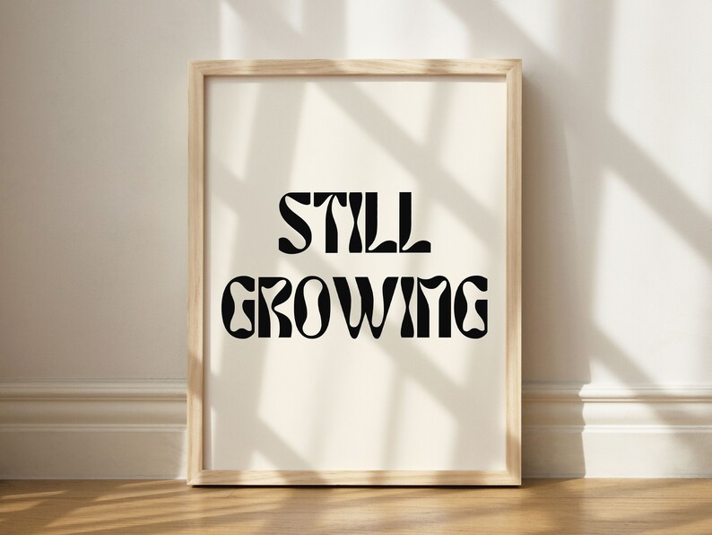 K&ouml;nnte beinhalten: Ein gerahmter Druck mit dem Text "STILL GROWING" in einer Retro-Schriftart. Der Druck ist auf wei&szlig;em Hintergrund und in einem hellen Holzrahmen gerahmt.