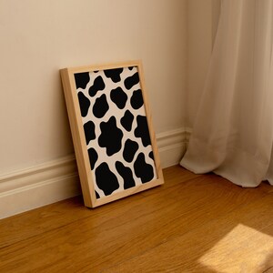 May include: A framed print with a black and white cow print pattern. The print is in a light wood frame and is leaning against a white wall.