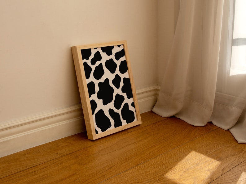 May include: A framed print with a black and white cow print pattern. The print is in a light wood frame and is leaning against a white wall.