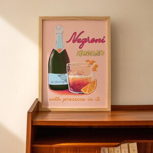 Pu&ograve; includere: Una stampa incorniciata con sfondo rosa che presenta un bicchiere di Negroni Sbagliato con prosecco, una bottiglia di prosecco e fette d'arancia. Il testo sulla stampa recita "Negroni Sbagliato with prosecco in it".