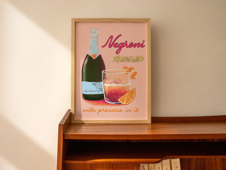 Pu&ograve; includere: Una stampa incorniciata con sfondo rosa che presenta un bicchiere di Negroni Sbagliato con prosecco, una bottiglia di prosecco e fette d'arancia. Il testo sulla stampa recita "Negroni Sbagliato with prosecco in it".