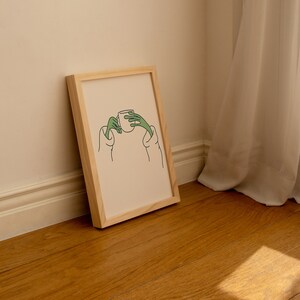 May include: A framed illustration of a person holding a cup with their hands. The illustration is in black and green line art on a cream background.