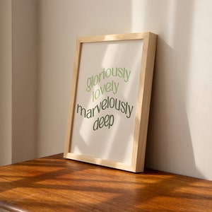 May include: A framed print with the text "gloriously lovely marvelously deep" in green lettering on a white background. The frame is made of light wood.