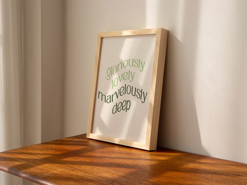 May include: A framed print with the text "gloriously lovely marvelously deep" in green lettering on a white background. The frame is made of light wood.