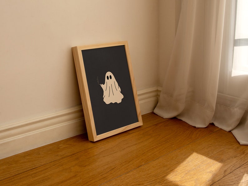 May include: A framed print with a black background and a white ghost illustration. The ghost is wearing a sheet and has two eyes. The frame is made of wood and has a light brown finish.
