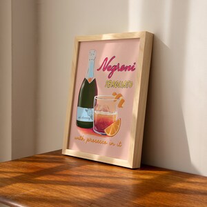 Pu&ograve; includere: Una stampa incorniciata con sfondo rosa che mostra un bicchiere di Negroni Sbagliato con prosecco, una bottiglia di prosecco e il testo "Negroni Sbagliato with prosecco in it".