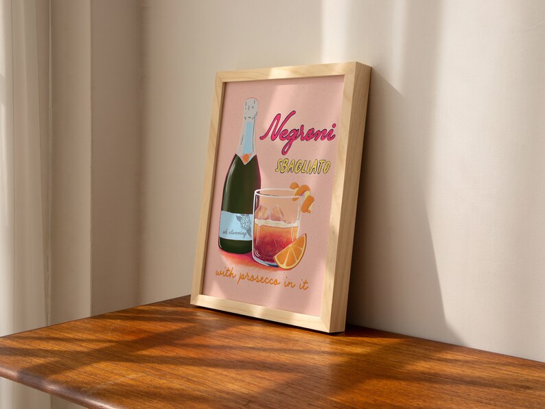 Pu&ograve; includere: Una stampa incorniciata con sfondo rosa che mostra un bicchiere di Negroni Sbagliato con prosecco, una bottiglia di prosecco e il testo "Negroni Sbagliato with prosecco in it".