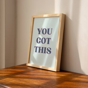 May include: A framed print with the text "YOU GOT THIS" in a light blue frame with a light wood frame.
