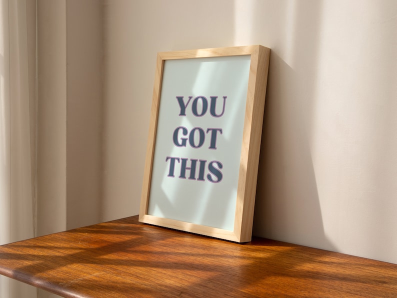 May include: A framed print with the text "YOU GOT THIS" in a light blue frame with a light wood frame.