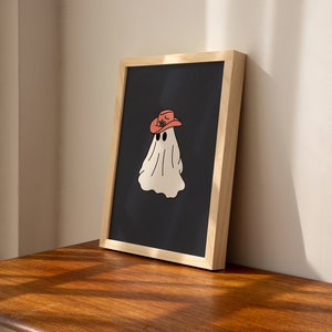 May include: A framed print of a white ghost wearing a red cowboy hat on a black background. The frame is made of light wood.