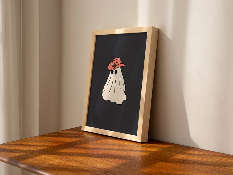 May include: A framed print of a white ghost wearing a red cowboy hat on a black background. The frame is made of light wood.