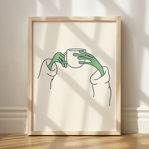 May include: A framed illustration of two green hands holding a white cup. The hands are drawn in a simple, cartoon style. The background is a light beige colour.